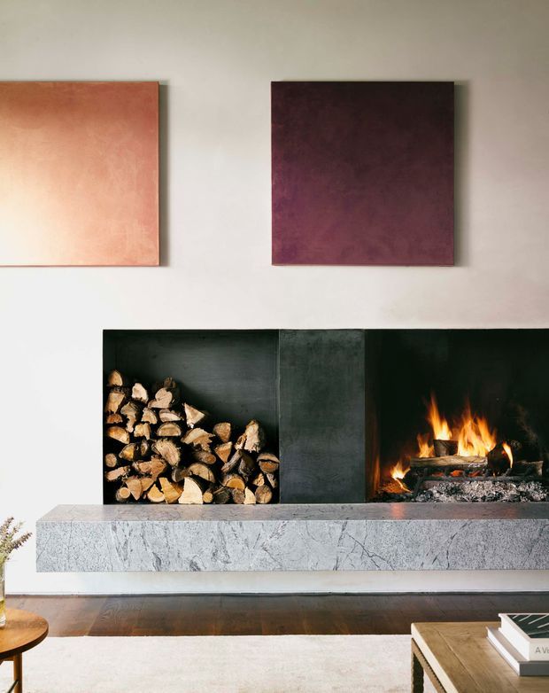 Interior view of a refined living room fireplace wall. A wide, dark built-in fireplace surround contains a burning open fire on the right and neatly stacked firewood on the left, with a substantial pale veined stone hearth. Two abstract square artworks hang above on a light wall, while partial timber furniture and a pale rug are visible in the foreground.