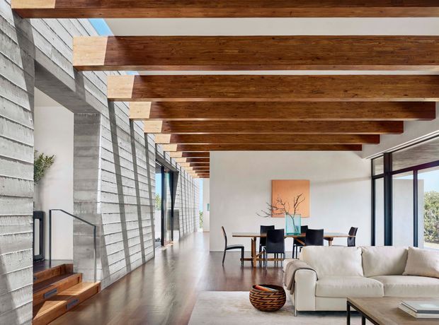 Open-plan contemporary living and dining space with a long linear layout, exposed dark timber ceiling beams, polished concrete block walls, timber flooring, expansive glazing, and a neutral sofa and dining setting. A timber stair rises along the left wall, while large windows and sliding glass doors bring in abundant daylight.