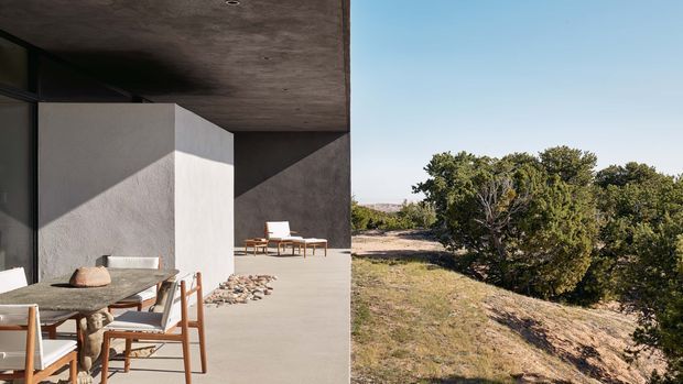 A minimalist modern concrete-covered outdoor terrace beside a dark glazed building, furnished with a timber dining table and chairs. The terrace opens toward a dry, shrub-covered natural landscape, with strong sunlight and sharp architectural shadows.