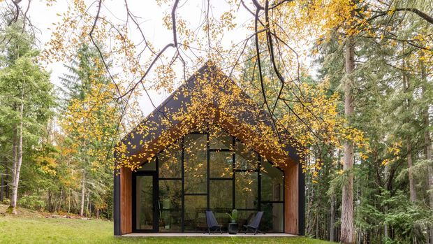 A small contemporary cabin or pavilion set within a dense forest, viewed from the front. The building has a steep gabled roof, dark exterior cladding, warm timber side walls, and a broad black-framed glazed facade. Autumn foliage partially frames the structure, with a simple outdoor seating area visible through the glazing.