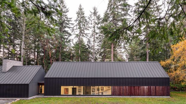 A low, modern single-storey building set within a dense forest. The dark standing-seam pitched roof and near-black vertical cladding create a restrained contemporary facade, with a long strip of warm-lit glazing and timber-toned cladding along the front. A smaller connected volume is visible on the left, and the building sits on a grassy clearing surrounded by tall trees.