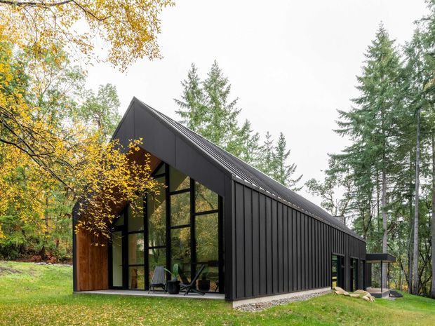 Exterior view of a contemporary, barn-like black-clad building set within a wooded landscape. The structure has a steep gabled roof, vertical dark metal cladding, extensive floor-to-ceiling glazing on the gable end, and a large timber entry door. A small outdoor seating area is visible at the glazed frontage.