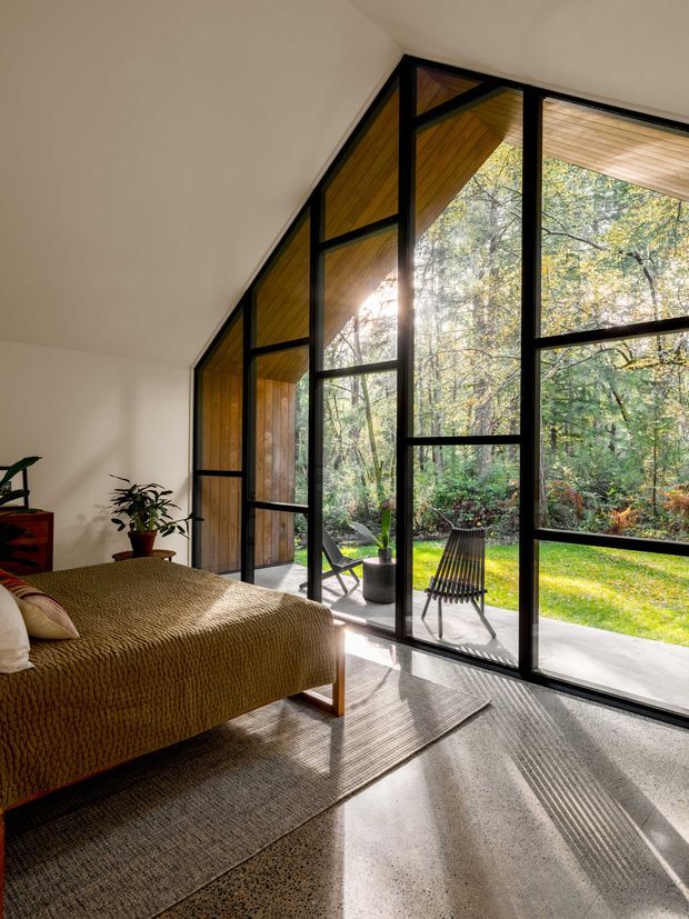 A warm, sunlit bedroom with a low sloped ceiling and a dramatic floor-to-ceiling glazed wall in black frames. The room includes a bed with a textured brown cover, a large neutral woven rug, indoor plants, and direct views onto a leafy garden and patio through the glazing. Exposed timber is visible in the window recesses and exterior overhang.