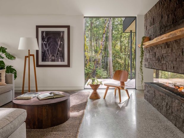 A refined contemporary living room with a polished concrete floor, neutral walls, a textured dark stone fireplace wall with timber mantel shelves and a linear fire, and a large floor-to-ceiling glazed opening overlooking a wooded setting. The room includes a low round timber coffee table, upholstered seating, a sculptural timber lounge chair, a floor lamp, framed artwork, and indoor plants. Natural light creates a warm, calm connection to the surrounding trees.