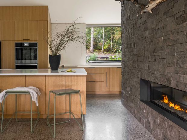 A contemporary kitchen with warm vertical-grain timber cabinetry, a white island benchtop, integrated black wall oven, and two slim metal-framed stools. A large rough-stacked dark stone fireplace with a long linear firebox dominates the right side, while a broad window provides a view to trees and landscaping. The polished grey floor and restrained furnishings create a warm, refined modern interior.