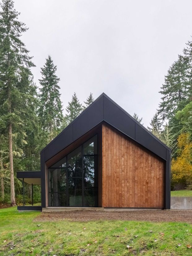 Exterior view of a compact modern gable-roof building set within a dense wooded landscape. The facade combines dark matte cladding and trim with warm vertical timber wall cladding, large black-framed glazed openings, and a projecting flat-roofed side element. The image has a restrained, contemporary woodland retreat character.