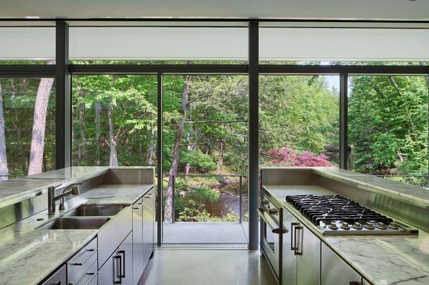 A contemporary kitchen viewed toward a wall of large, dark-framed windows and a central glazed door overlooking dense woodland. Two parallel runs of pale cabinetry and stone benchtops frame the view, with a sink and tap on the left, a large gas cooktop on the right, and streamlined modern hardware. The space is bright, refined, and strongly connected to the surrounding landscape.