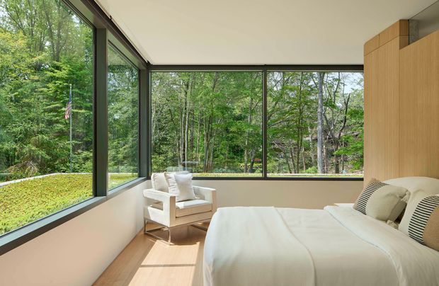Bright, compact bedroom with floor-to-ceiling corner glazing overlooking a wooded landscape. The room features a low-profile bed with white bedding, a light upholstered lounge chair, pale timber flooring, a warm light-wood built-in wall or headboard, and slim dark window frames. The clean-lined interior has a calm, contemporary feel and strong indoor-outdoor connection.