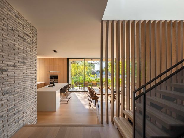 A warm contemporary open-plan interior viewed from a stair landing, with a pale exposed brick feature wall, timber kitchen cabinetry and island, dining area, full-height glazing, and a timber vertical-slat screen beside a black metal stair balustrade. Natural light and garden views create an airy connection to the outdoors.