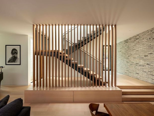 A warm, contemporary interior focused on a timber staircase screened by closely spaced vertical timber slats. The stair rises behind built-in timber cabinetry and leads toward an upper level, with a textured pale brick feature wall to the right, light timber flooring, and a dining area in the foreground. Neutral walls, integrated joinery, and restrained furnishings create a refined, architectural composition.