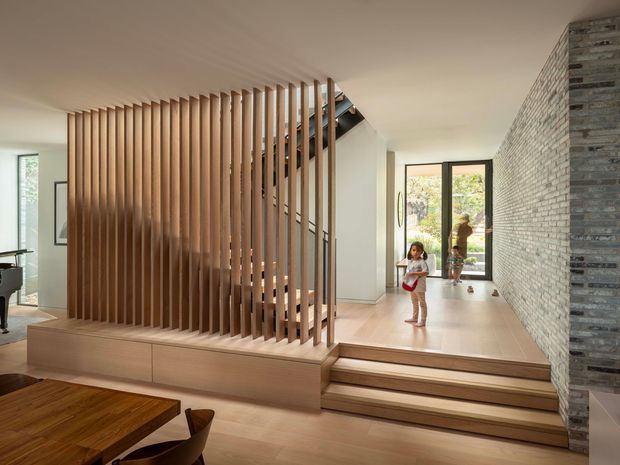Open-plan contemporary residential interior featuring a timber dining area in the foreground, a sculptural staircase screened by vertical timber slats, pale timber flooring and built-in joinery, and a textured grey stone-clad wall. Large windows and glazed doors bring in natural light, with a small garden courtyard visible beyond.