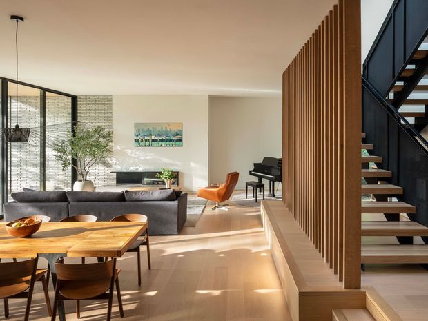 A bright, open-plan contemporary interior combining a dining area, living room, and stairway. The space features pale timber flooring, a large timber dining table with curved wooden chairs, a charcoal sectional sofa, a brick feature wall, full-height black-framed glazing, a vertical timber slat screen with integrated bench seating, and an open-riser staircase with dark metal structure. Natural light creates a warm, refined atmosphere.