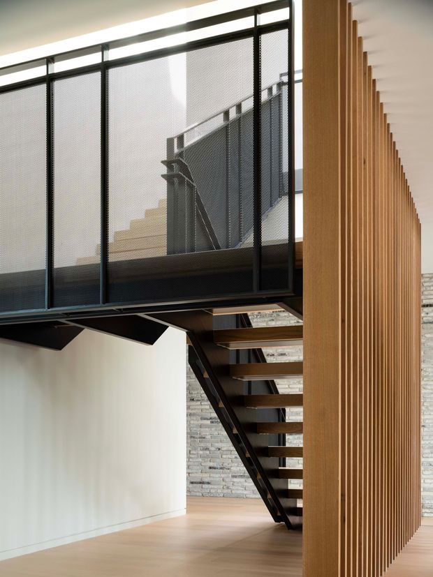 Interior architectural view of a contemporary stairway and mezzanine. Open-riser timber treads are supported by a dark metal structure, with a black perforated-metal balustrade enclosing the upper-level walkway. A tall vertical timber slat screen runs alongside the stairs, while a pale exposed brick wall and light timber floor add texture and warmth. The space is bright, minimalist, and strongly defined by layered vertical and diagonal lines.