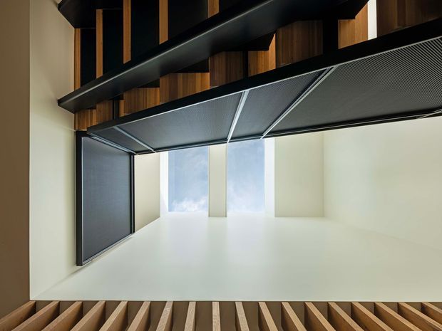 Upward-looking architectural interior view focused on a dramatic double-height void. Dark exposed structural beams and warm timber battens frame a bright white space, with tall vertical glazing or skylight openings bringing in natural light. A perforated or finely patterned dark ceiling plane spans part of the opening, creating a strong contrast between black, timber, and white surfaces.