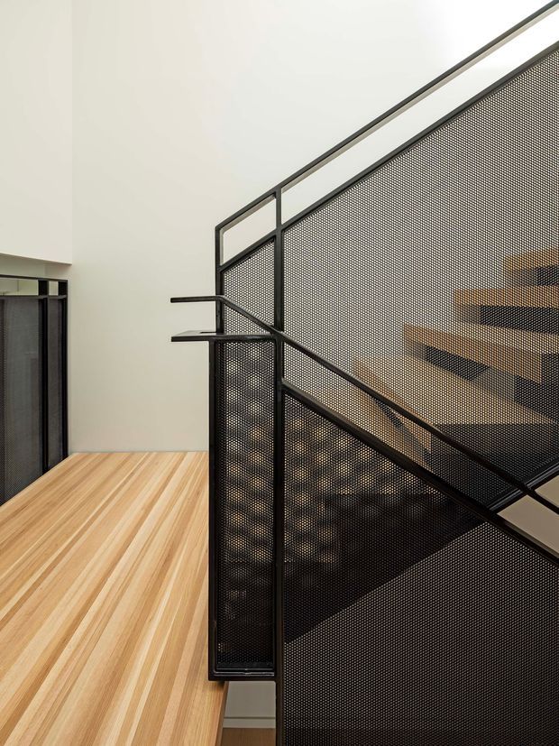 Interior view of a contemporary stairway with a black perforated metal screen and matching steel handrail forming a sculptural balustrade. Warm timber flooring and stair treads contrast with the dark metalwork, while a tall white wall and glimpses of the stair structure create a clean, minimalist composition.