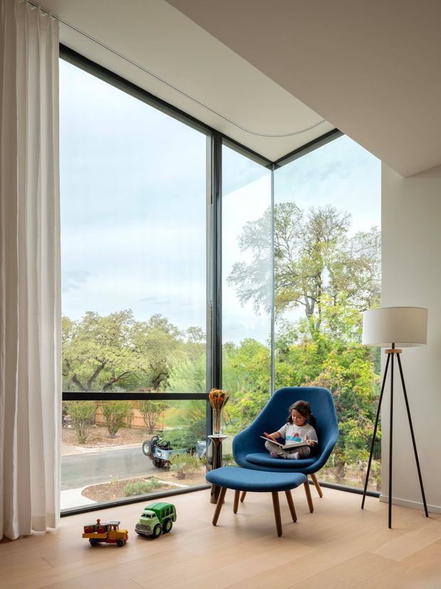 Bright, minimalist interior corner with floor-to-ceiling corner glazing, pale timber flooring, white walls, and sheer curtains. A deep blue mid-century style lounge chair, tripod floor lamp, child reading, and toys create a calm family living space overlooking leafy suburban surroundings.