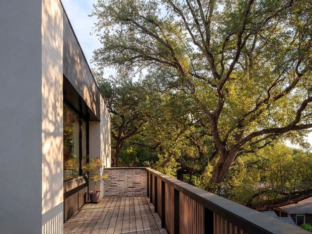 A contemporary upper-level outdoor deck or balcony beside a minimalist building facade, surrounded by mature leafy trees. The deck has timber flooring, a dark timber or metal balustrade, and a low brick feature wall near the building. Large windows open toward the deck, with warm filtered daylight creating a tranquil connection to the surrounding landscape.