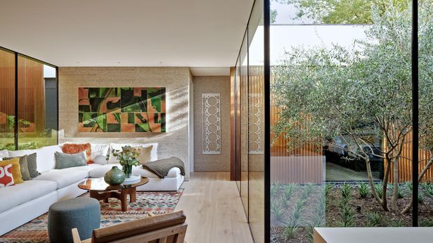 A refined contemporary living room opening onto a landscaped courtyard through extensive floor-to-ceiling glazing. The space features a large white sectional sofa, sculptural timber coffee table, upholstered ottoman, patterned rug, textured light stone or masonry walls, warm timber screening, and a colourful abstract artwork. The courtyard includes mature planting and timber fencing, creating a calm indoor-outdoor connection.