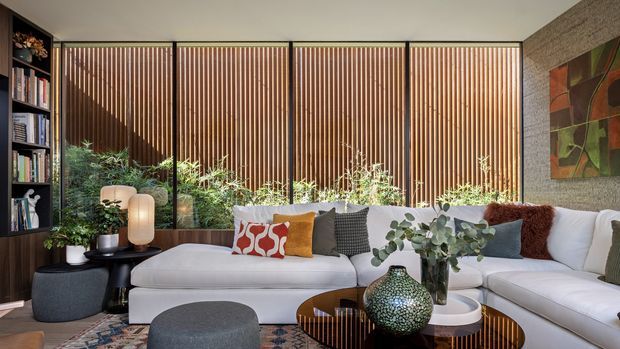 A refined contemporary living room with a large white sectional sofa arranged around layered round coffee tables and upholstered ottomans. Full-height glazing is screened by warm vertical timber battens, with indoor plants, open shelving, textured cushions, a large decorative vase, and a colourful artwork contributing to the styling. The room has a warm, sophisticated, indoor-outdoor character with strong natural light.