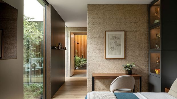 A refined residential interior showing a compact study or writing nook beside a bedroom or living area. The space features textured beige wall covering, dark timber or painted built-in cabinetry, a slim timber console desk, upholstered chair, warm timber flooring, framed artwork, and full-height glazing overlooking greenery. The palette is warm and earthy with carefully integrated joinery and soft natural light.