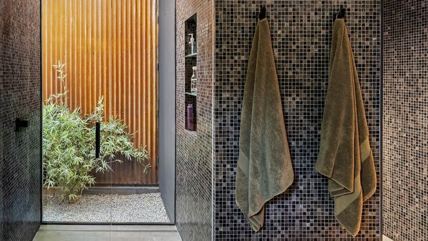 A contemporary bathroom interior with dark small-format mosaic tile walls, a frameless glazed opening, and warm vertical timber screening beyond. Two olive-brown towels hang from simple black hooks, while a planted gravel lightwell adds natural texture and daylight. The room has a refined, earthy, spa-like atmosphere.