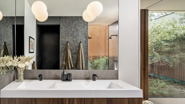 A contemporary bathroom vanity with a wide double-sink white benchtop, dark timber cabinetry, black tapware, and a large wall-to-wall mirror. The room features a dark small-format mosaic tile wall, pale painted walls, sculptural globe pendant lights, wall-mounted towel hooks, and a glazed shower area with a vertical timber screen. Garden greenery is visible through the full-height window.