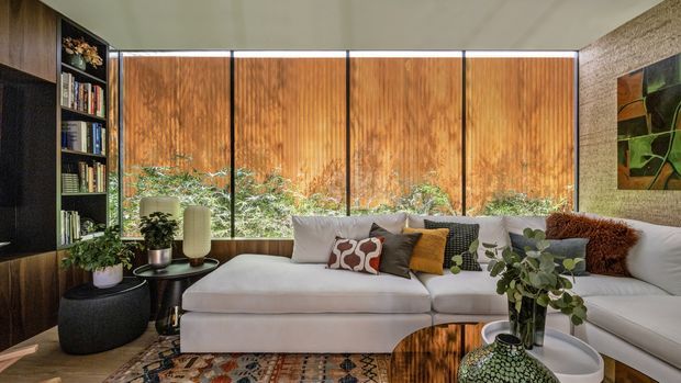 A warm, richly layered living room with a large L-shaped white sectional sofa, patterned and textured cushions, a round dark coffee table, indoor plants, bookshelves, dark built-in joinery, and full-height glazing screened by timber-toned vertical panels. A textured feature wall and colourful artwork add depth, while a patterned rug and polished wood table reinforce the inviting, eclectic atmosphere.