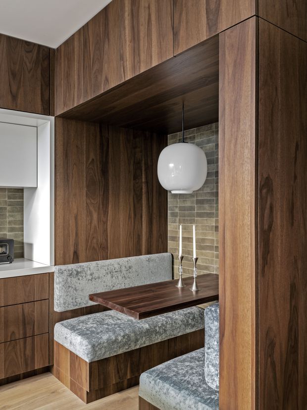 A compact built-in dining nook beside a kitchen, enclosed by rich dark timber wall panelling and joinery. The nook has upholstered bench seating, a dark timber dining table, a white globe pendant light, and a muted grey-green rectangular tiled backsplash. Light timber flooring and white kitchen cabinetry are visible at the left.