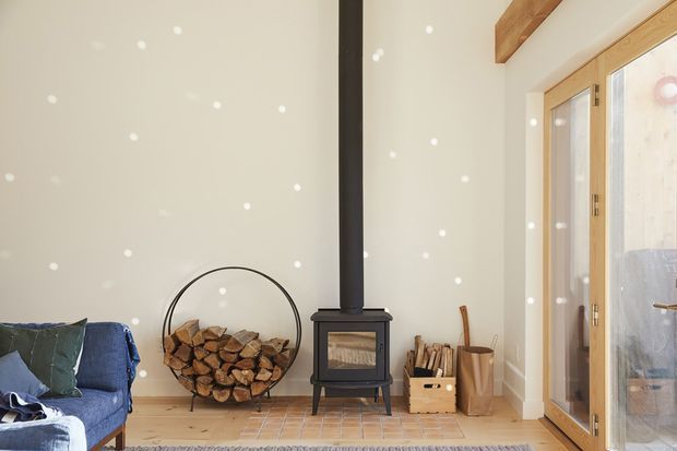 Bright, cozy living room with a freestanding black wood-burning stove and exposed flue, a circular firewood holder filled with logs, pale timber flooring, light walls, exposed timber ceiling beams, and large timber-framed glazed doors. A blue sofa is partially visible at left, with a calm natural material palette and strong indoor-outdoor connection.