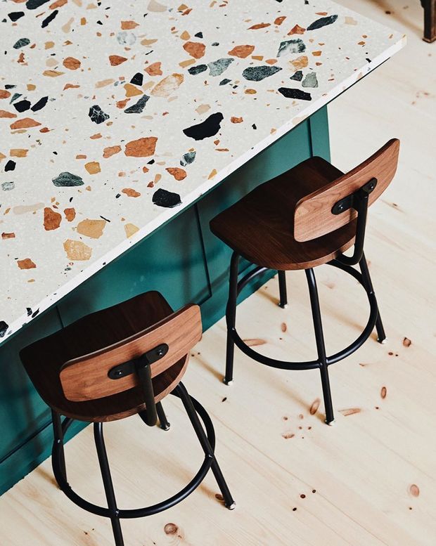 Close-up interior view of a compact dining or kitchen counter area with a white terrazzo benchtop containing multicoloured stone chips, set over teal cabinetry or a panelled base. Two dark wood and black metal bar stools sit beneath the counter. The space has pale timber plank flooring and a bold, playful contemporary colour palette.