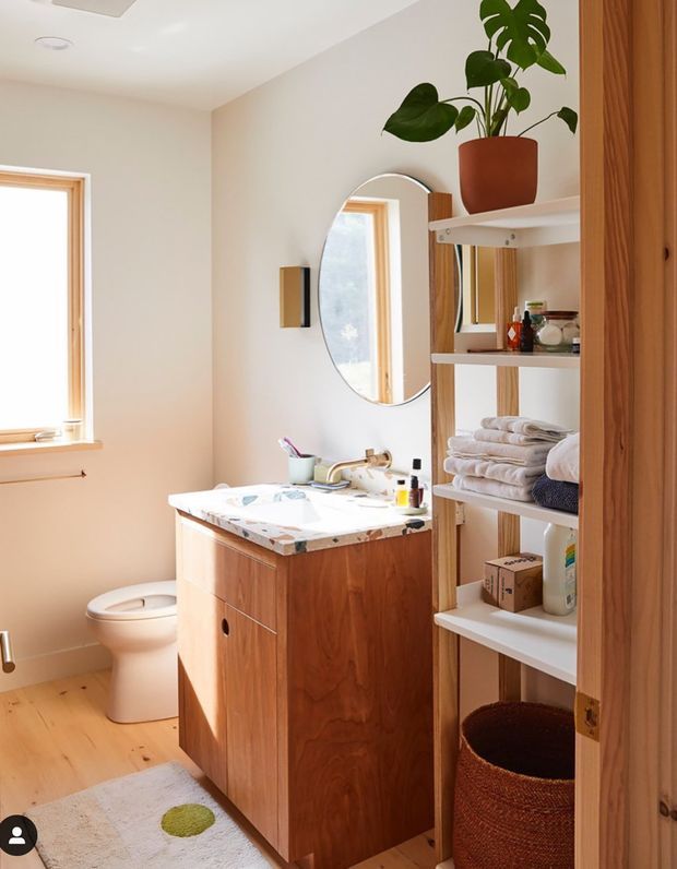 Bright, compact bathroom with a timber vanity, pale stone-patterned benchtop, wall-mounted basin, oval mirror, toilet, timber flooring, and open white shelving stocked with towels and toiletries. Warm natural light enters through a timber-framed window, while a potted monstera adds greenery. The room has a clean, warm, natural contemporary feel.