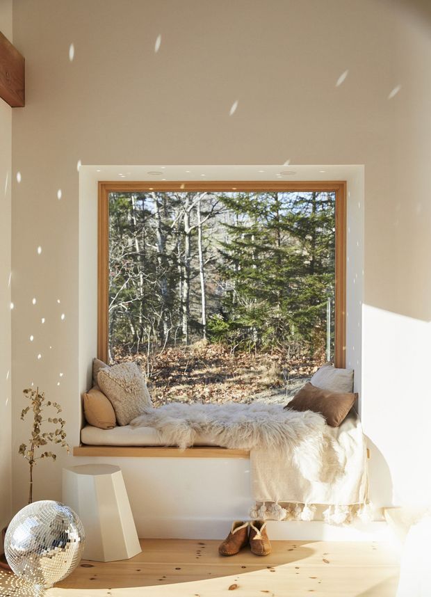 A bright, serene window nook with a deep timber-framed picture window overlooking a wooded landscape. The built-in timber window seat is layered with neutral cushions, a fluffy sheepskin throw, and a cream blanket. Pale walls, warm timber flooring, a faceted white side table, disco ball, and soft sunlight create a cozy, Scandinavian-inspired retreat.