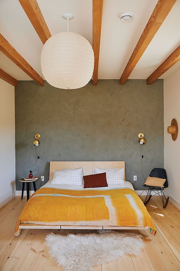 A warm, compact bedroom with a low platform bed centered against a textured grey feature wall. The room has exposed timber ceiling beams, pale timber flooring, a mustard and white striped bedspread, simple grid-pattern bedding, wall-mounted sconces, a suspended white paper pendant, and a black accent chair. A white sheepskin rug adds softness in the foreground.