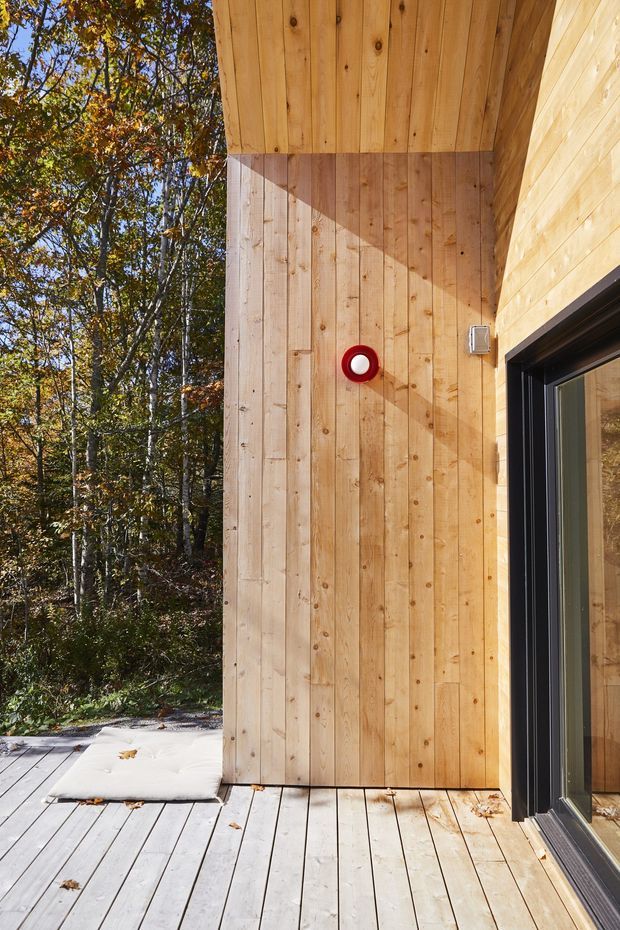 Close-up exterior view of a timber-clad building at the edge of a wooded site. The image shows vertical natural wood wall boards, a matching timber soffit, a pale timber deck, and a large dark-framed glazed door. A small red-and-white circular wall light provides a vivid accent. Forest vegetation is visible beyond the deck.