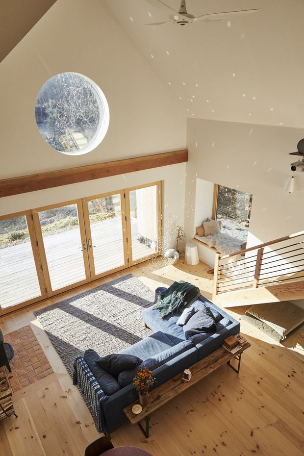 A bright, airy double-height living room viewed from an upper level, featuring a large blue sectional sofa, timber flooring, a long timber coffee table, a textured area rug, exposed timber beam, expansive timber-framed glazed doors, a round porthole-style window, and a stair with slim black metal balustrade. Sunlight creates dappled reflections across the white walls and floor, and a window seat occupies the far side of the room.