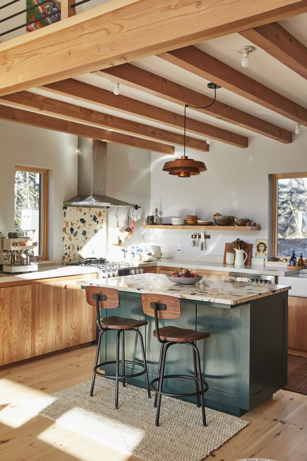 A warm, light-filled kitchen with exposed timber ceiling beams and a large central island. The island has a mottled stone benchtop and dark green fixed cabinetry, paired with two timber-and-black-metal stools. Timber cabinetry, open shelving, a stainless-steel rangehood, terrazzo-style splashback, pendant lighting, and timber flooring define the space.