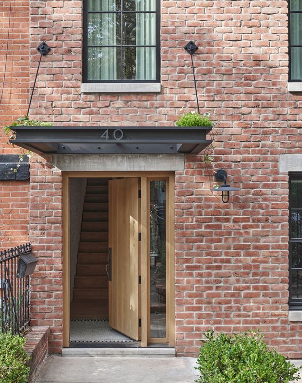 Close-up exterior view of a brick townhouse entrance. The facade features red brick masonry, black-framed multi-pane windows, a timber pivot entry door with a narrow glazed sidelight, black wall-mounted lanterns, planters, and a projecting metal-and-concrete entry canopy marked with the number 40.
