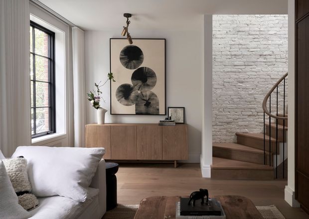 A refined living room with a white sofa in the foreground, pale timber flooring, and a low mid-century-style timber sideboard against a light wall. A large abstract monochrome artwork hangs above the sideboard, with a sculptural pendant light overhead. Black-framed windows with sheer curtains bring in natural light on the left, while a white-painted exposed brick wall and curved timber staircase define the adjoining area on the right. The room has a warm, restrained contemporary aesthetic with subtle mid-century influences.