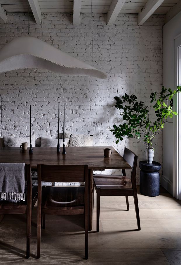 A moody, refined dining area with a dark timber dining table and chairs, set against a white-painted exposed brick wall. White-painted ceiling beams, pale timber flooring, black taper candleholders, and a tall leafy plant in a sculptural dark planter create a warm, textural interior. A large abstract white sculptural artwork hangs above the table, with daylight entering from the right.
