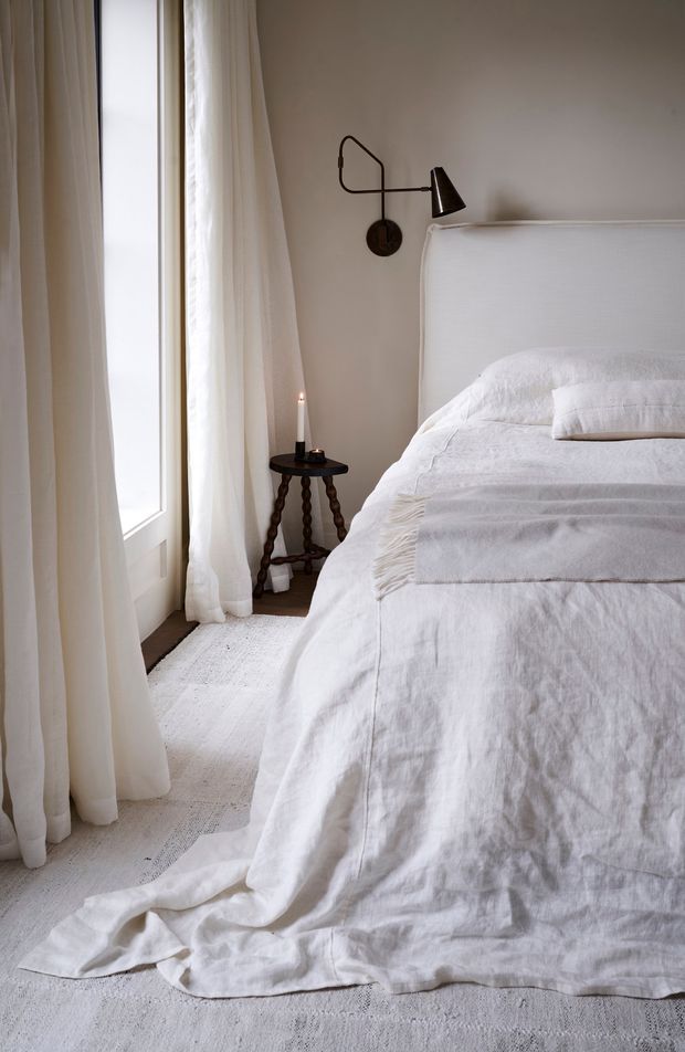 A softly lit, calm bedroom corner featuring a large upholstered white bed with layered white linen bedding, a textured white area rug, full-height sheer curtains beside a tall window, and a dark sculptural bedside table with a candle. A black wall-mounted reading lamp provides contrast against the warm off-white wall.