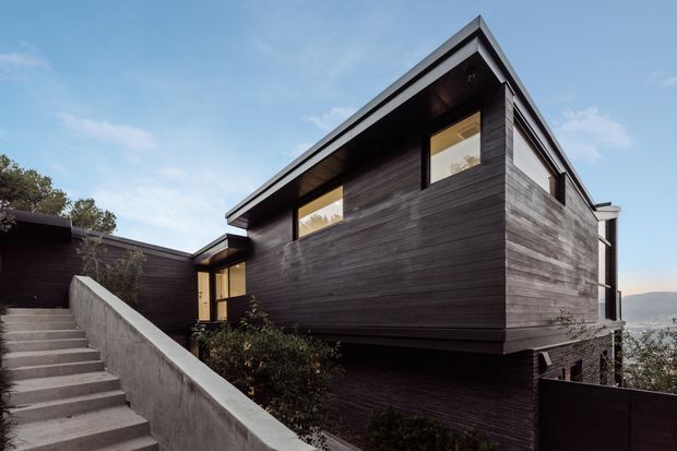 Exterior view of a contemporary multi-level home on a sloping site, featuring dark horizontal timber cladding, sharply angled rooflines, recessed windows, and concrete entry stairs. Water and distant landscape are visible beyond the building.