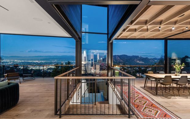 A sophisticated open-plan upper-level interior with dramatic floor-to-ceiling glazing overlooking a city basin and mountains at dusk. The space combines a dining area with a large timber table and upholstered chairs, a relaxed lounge area, and a central stair void enclosed by slim black metal balustrades. Exposed dark structural beams and a coffered timber ceiling create a strong contemporary architectural character, while warm timber flooring, a patterned rug, and indoor planting soften the space.