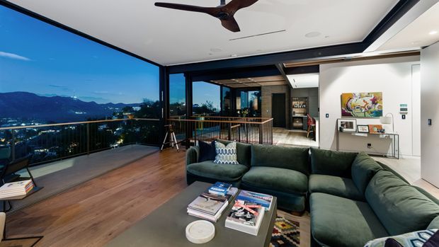 A contemporary open-plan living room at dusk with a large dark green sectional sofa, coffee table, timber flooring, and extensive floor-to-ceiling glazing opening onto a balcony with a distant mountain and city view. The room includes a ceiling fan, recessed lighting, a compact console display area, and a visible adjoining kitchen or bar zone beyond the railing.