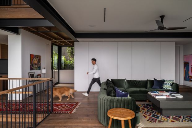 A contemporary open-plan living room viewed from an adjoining stair landing. The space features a large deep green sectional sofa, dark rectangular coffee table, patterned rugs, timber flooring, white full-height built-in wall panels, and exposed timber and dark structural ceiling elements. A black metal balustrade borders the stair opening in the foreground, with framed artwork, indoor planting, and a ceiling fan adding detail.