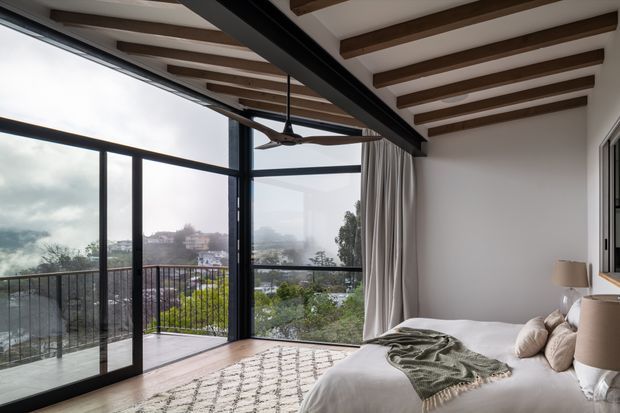 A bright contemporary bedroom with a large corner of black-framed floor-to-ceiling glazing opening onto a balcony and a leafy hillside view. The room features a white bed with neutral bedding, bedside lamps, full-height curtains, pale timber flooring, and a white ceiling with exposed dark timber beams and a ceiling fan.