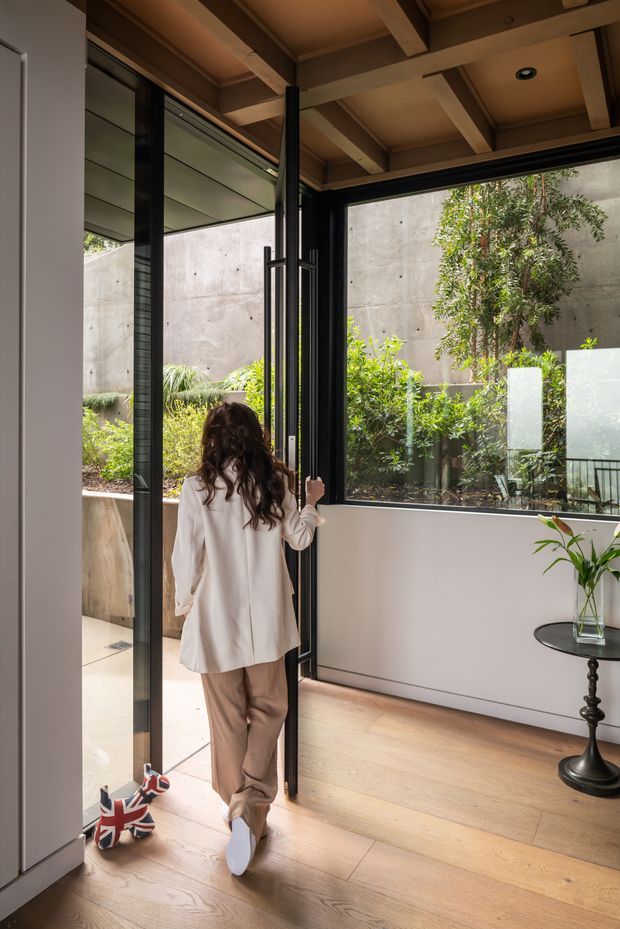A warm contemporary interior opening onto a landscaped courtyard. The room features wide light timber flooring, large black-framed glazing and a full-height glazed door, exposed timber ceiling beams, pale walls, and a slim black pedestal side table with a vase. A person is opening the door, emphasizing the connection between the interior and the planted outdoor space.