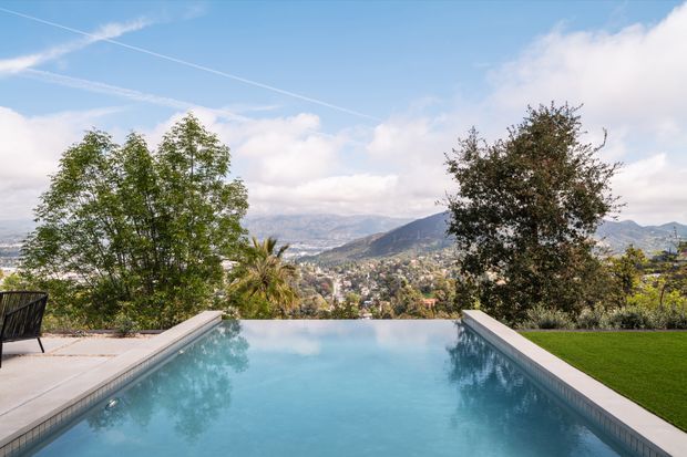 An outdoor infinity-edge swimming pool occupies the foreground, with pale paving around it and a manicured lawn to the right. Trees frame a broad elevated view over a hilly, settled landscape beneath a bright blue and partly cloudy sky.