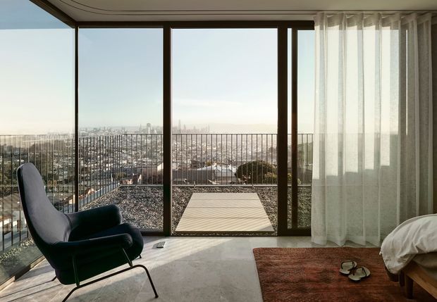 A bright, elevated interior seating area with floor-to-ceiling glazing opening onto a balcony terrace. The room features a sculptural dark upholstered lounge chair, sheer curtains, pale flooring, and a partially visible bed, with expansive urban views beyond the balustrade.