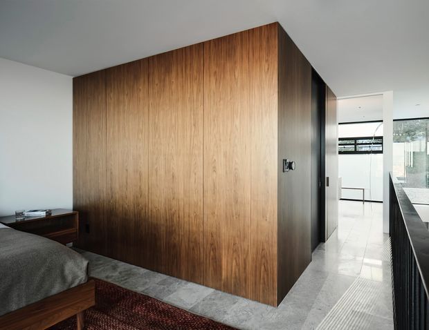A refined contemporary bedroom with a large freestanding timber-clad wardrobe or storage volume forming the central architectural feature. The room has a low timber bedside unit, upholstered bed, dark textured rug, pale stone or tile flooring, white walls and ceiling, and a bright corridor or bathroom-like space visible beyond. Black-framed glazing and dark linear detailing add contrast.