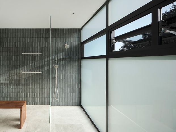 A minimalist contemporary bathroom with a walk-in glass shower, wall-mounted rainfall shower and hand shower, pale stone-like flooring, and a timber bath bench or vanity edge in the foreground. The bathroom is enclosed by slim black-framed glazing, including translucent lower panels and clear upper clerestory-style windows that bring in natural light and reveal trees outside. The wall behind the shower is finished in narrow vertically patterned grey tiles.